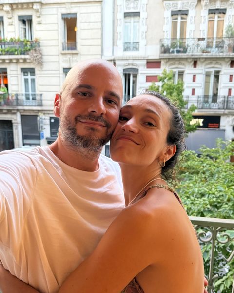 Juan and Pau cheek to cheek at an open window above a city street