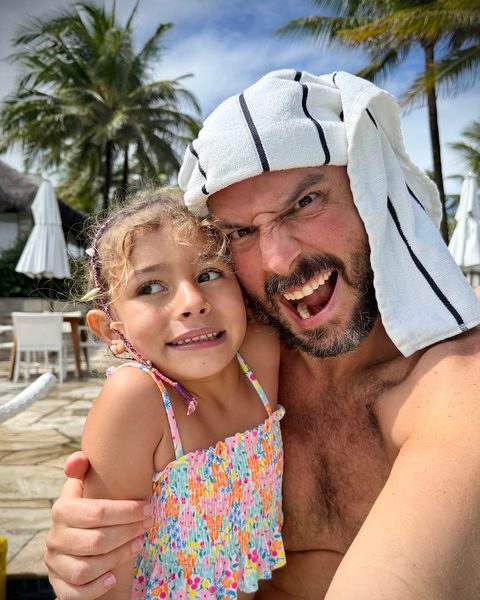 Juan, a towel on his head, pulling a fierce face beside a smiling Fran by a pool with palm trees
